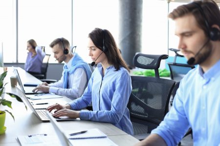 Smiling male call-center operator with headphones sitting at modern office with collegues on the backgroung, consulting online